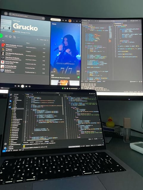 Developer workspace with laptop showing code editor and desktop monitors displaying Grucko app interface and programming code