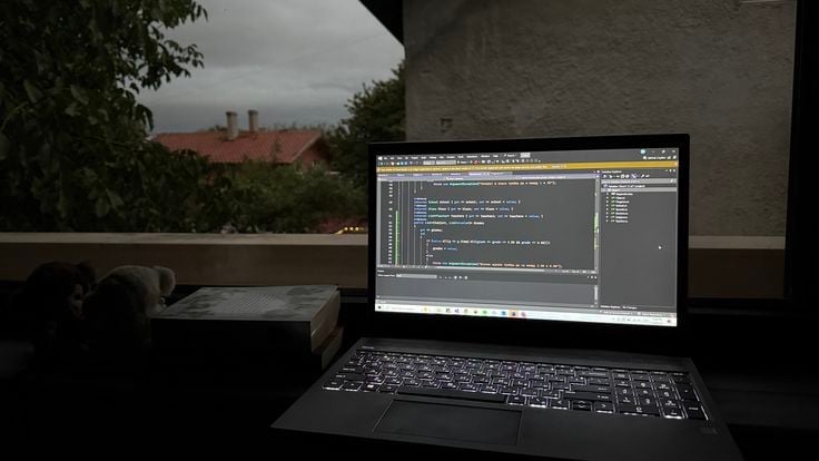 Laptop displaying code editor with programming interface, placed on desk near window with urban rooftop view