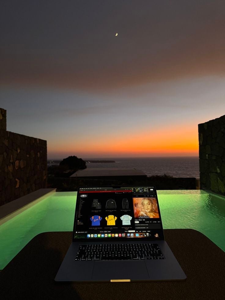 Laptop with design software displayed on glowing green surface overlooking sunset ocean with silhouetted buildings
