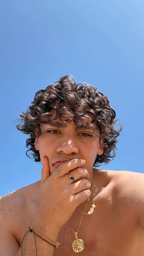 Person with curly dark hair touching their face against a clear blue sky, wearing jewelry and appearing to be at the beach